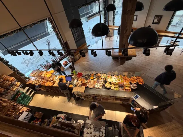 An overhead view of a long bakery counter with staff, black lamps and tall windows