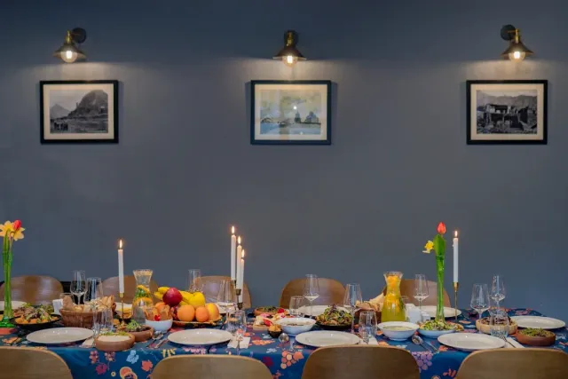 A long table in a blue floral cloth with candles, fruit and glasses, facing a wall of photographs
