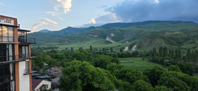 A balcony view over a green valley and mountain ridges, beside a brick building with balconies