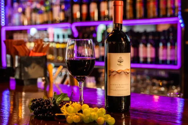 A bottle of red wine and a full glass on a counter, with bunches of grapes and purple lighting