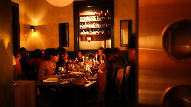 A dim candlelit space with diners at a long table and a lit shelf of bottles