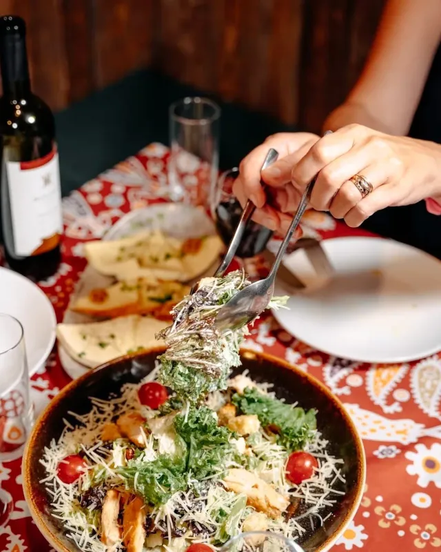 A hand lifting leaf salad with a fork from a bowl, on a printed red cloth beside a wine bottle