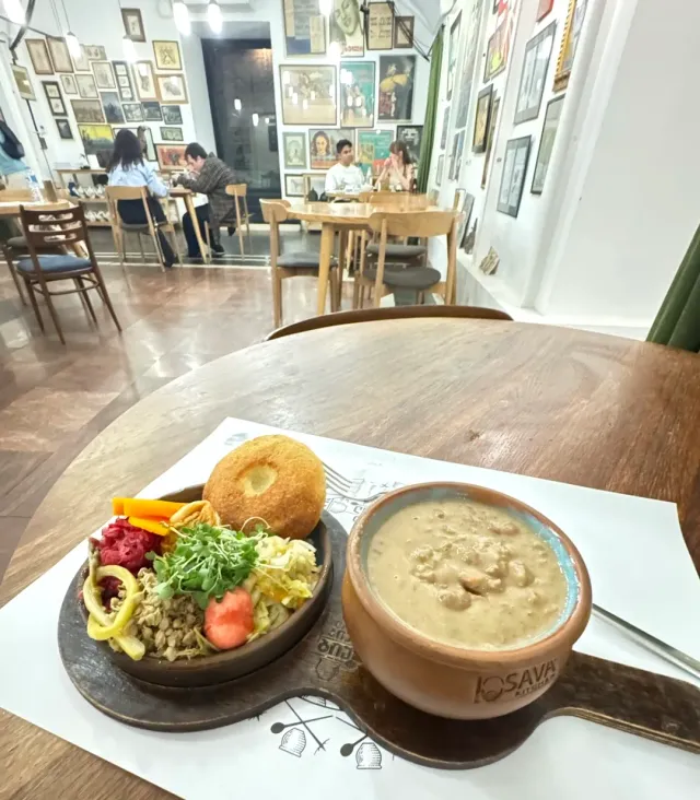 A clay pot with bean stew and a plate of accompaniments, against a wall of framed pictures
