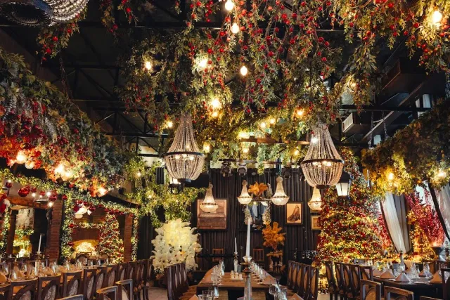A ceiling laden with greenery and red flowers with crystal chandeliers, and set tables beneath