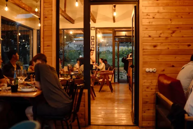 A wooden space lit in the evening with diners at tables, hanging bulbs and another room beyond a doorway