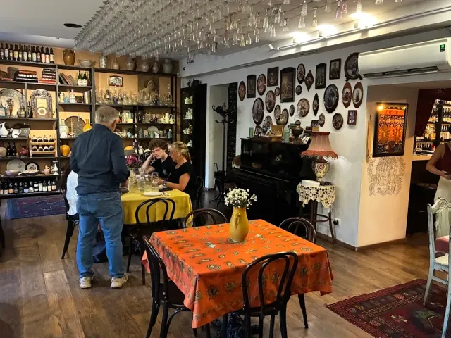A space with shelves of bottles, glasses hanging from the ceiling, a wall of plates and a black piano