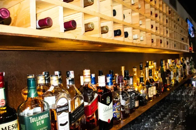 A long shelf of liquor bottles beneath a wooden wine rack, with glasses at the end of the counter