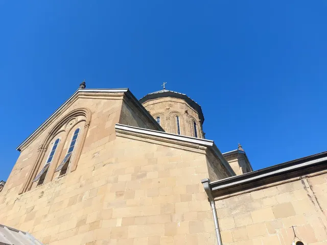 The stone facade of a church with arched windows and a dome behind under a blue sky