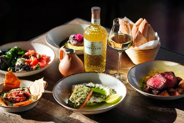A wooden table laden with salad, a fish fillet, slices of meat, a bread basket and a wine bottle