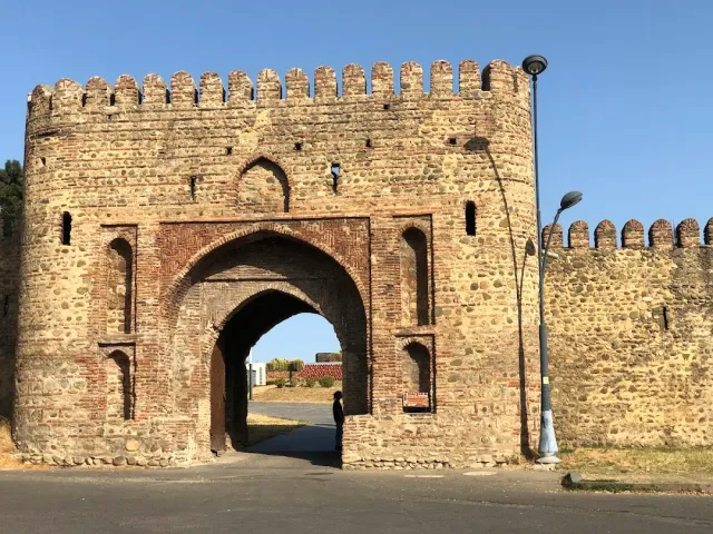 A stone fortress gate with a crenellated wall, a pointed brick arch and a person standing in the passage