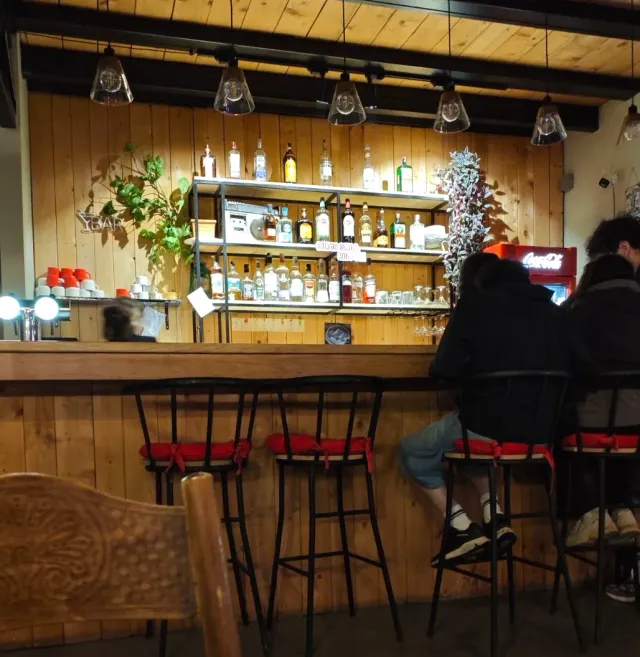 A wooden counter with bottle shelves, tall stools in red upholstery and people sitting to the side
