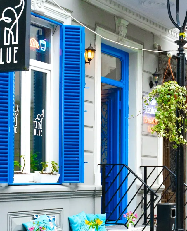 A white facade with bright blue shutters and door, a hanging sign, a trailing plant and turquoise cushions