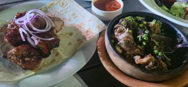 Slices of grilled meat on flatbread with onion rings, beside a clay pan and red sauce