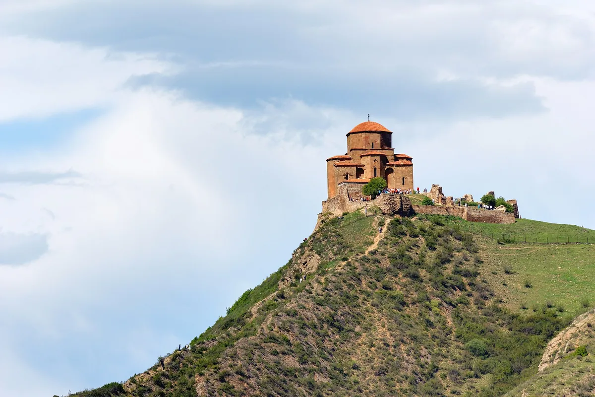 A stone church with a red dome on top of a green ridge against a cloudy sky