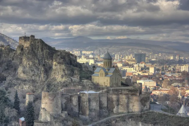The walls and towers of a stone fortress on a ridge above the city roofs