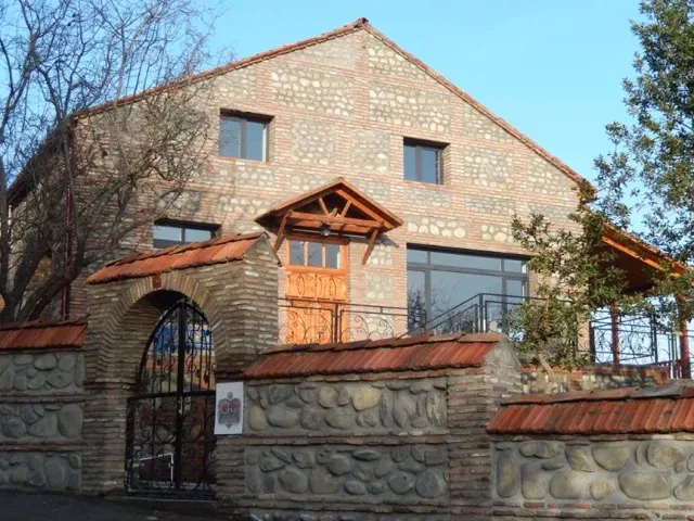 A stone and brick building with a tiled roof, a covered wooden entrance and an arched gate in a river stone wall