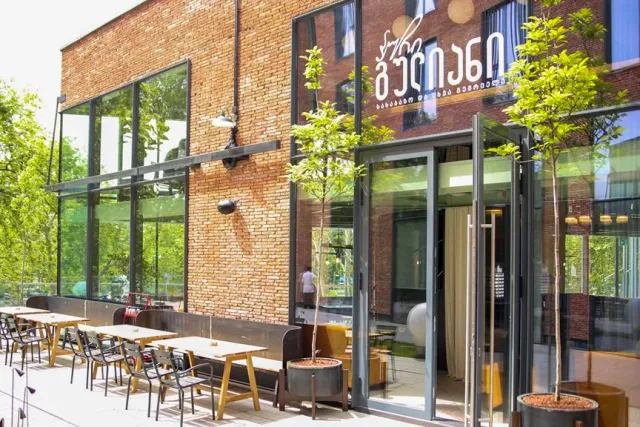 A brick building with large glass windows, wooden tables outside and young trees in planters