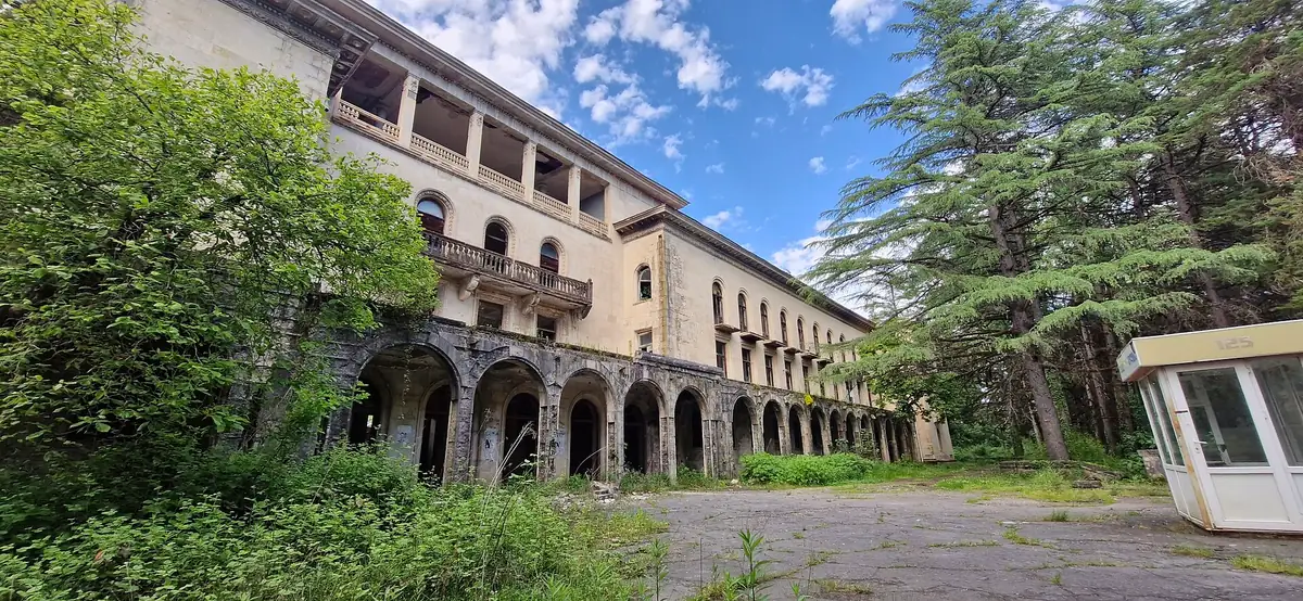 An abandoned building with rows of arches and balconies, weeds spreading across the forecourt