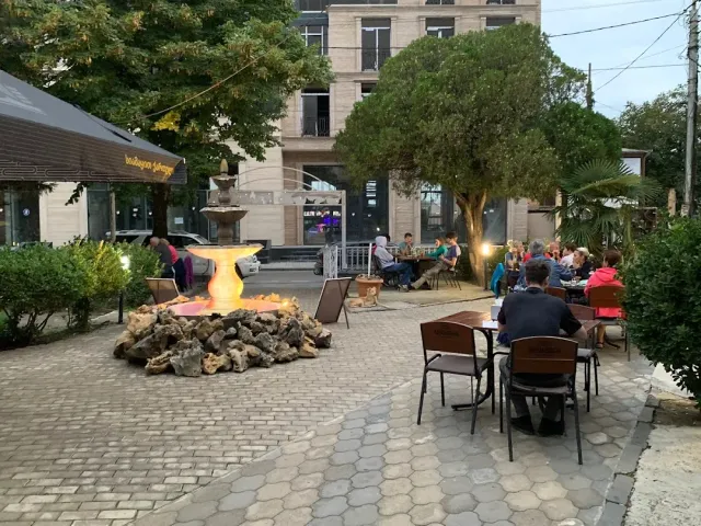 Tables on a paved plaza with a lit fountain and diners under trees in the evening