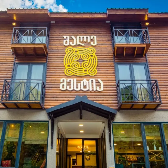 A two storey wooden facade with lettering and a yellow ornament, balconies and a lit glass entrance