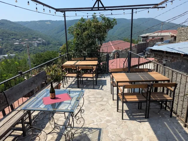 A rooftop terrace with wooden tables, a string of bulbs and a wine bottle, with green hills behind