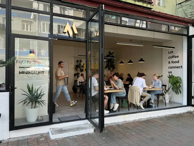 A glass front open to the street with guests around wooden tables and plants inside