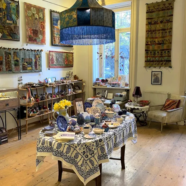 A gallery room with a table holding ceramics, woven wall hangings and a blue fringed lampshade
