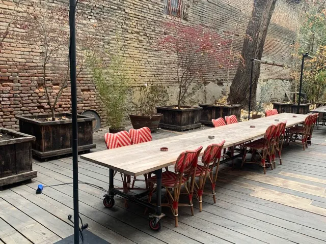 A long wooden table on a deck floor in a courtyard, red cane chairs and a brick wall