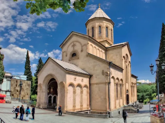 A stone church with a conical dome and arched decorations, and people on the plaza in front of it