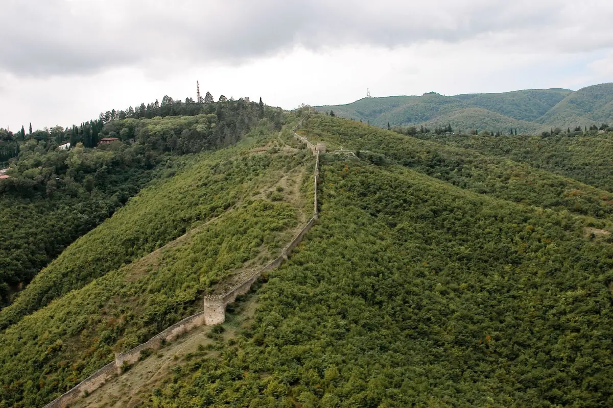 An old city wall climbing along a green wooded ridge, with a watchtower on the slope