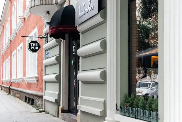 A pale street facade with a round hanging sign, a dark awning and a display window with plants