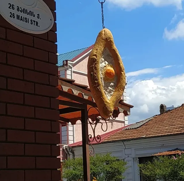 A hanging sign shaped like a boat bread with a yolk, against houses and a blue sky