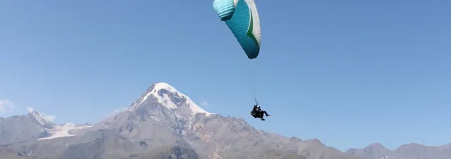 A turquoise paraglider carrying a tandem rider in front of a snow covered peak