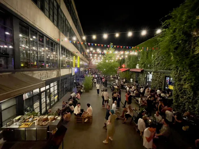 A busy courtyard in the evening with strings of lights, tables and a large crowd
