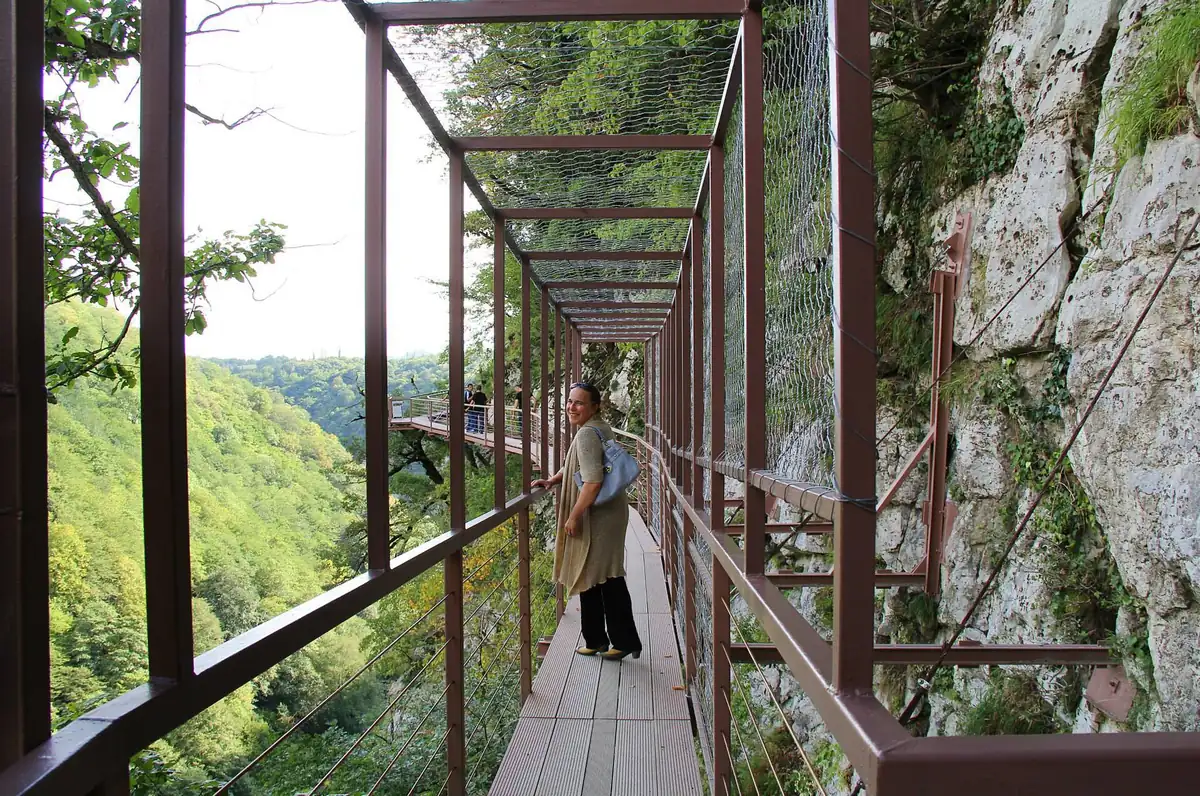 A metal walkway clinging to a cliff wall, with a hiker walking along it above a wooded valley