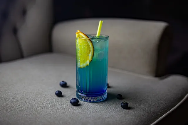 A blue cocktail with an orange slice and a yellow straw on an armchair, with blueberries around it