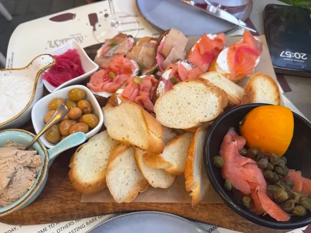 A wooden platter with baguette slices, smoked fish, olives, pickled onion and spread in small bowls