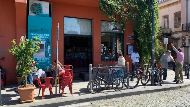 An orange facade with red chairs on the sidewalk, parked bicycles and passers by