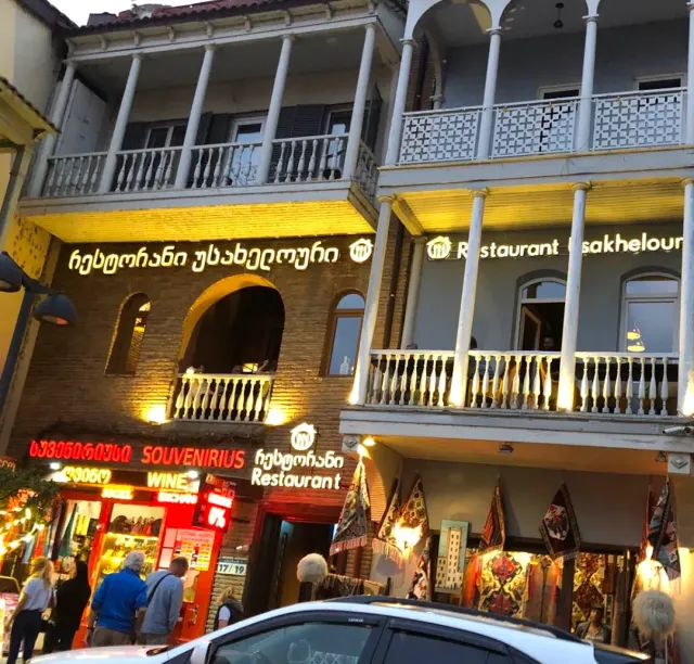 A building with columned balconies and lit signage in the evening, and a shop hung with carpets below