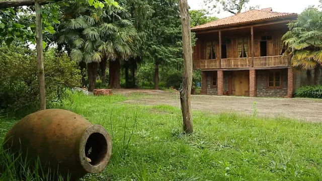 A large clay jar lying on the grass, with a two story wooden house with a balcony among trees behind