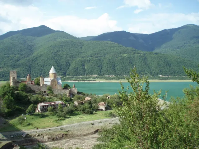 A walled fortress with a church on a green hill above a turquoise reservoir