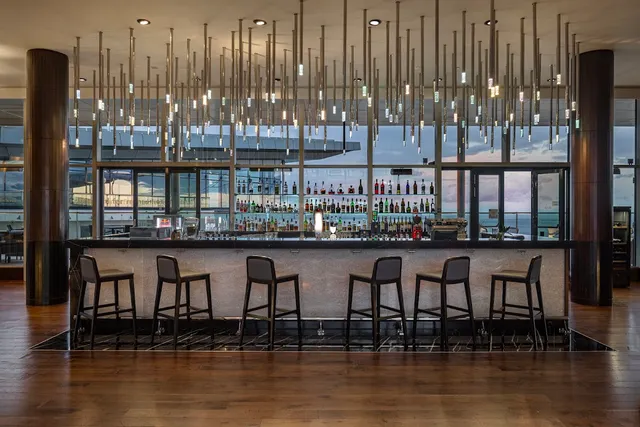 A long bar counter with high stools, shelves of bottles and hanging lights above