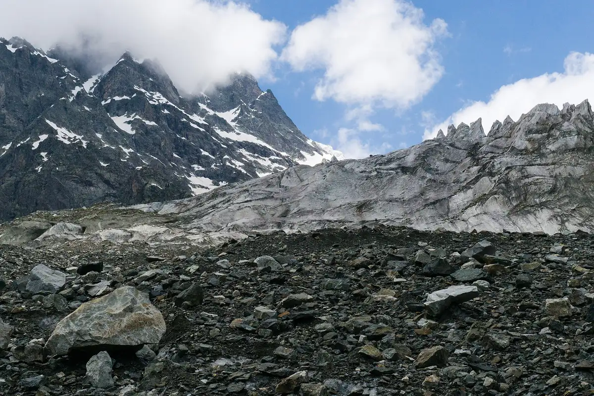 A grey glacier tongue and moraine boulders below jagged snow streaked peaks