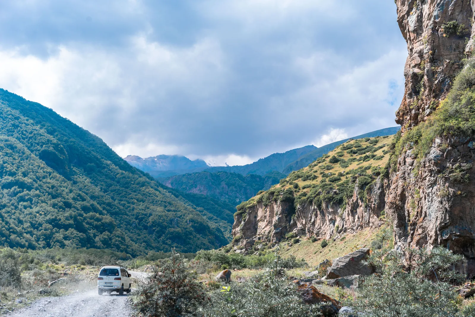 A road running below high rock walls through a narrow Caucasus gorge, a vehicle small against the cliff
